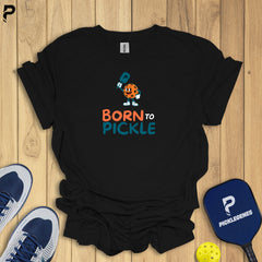 Black t-shirt with 'Born to Pickle' design on a wooden surface with pickleball equipment.