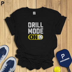 Black t-shirt with 'DRILL MODE ON' text on a wooden surface with sports equipment.