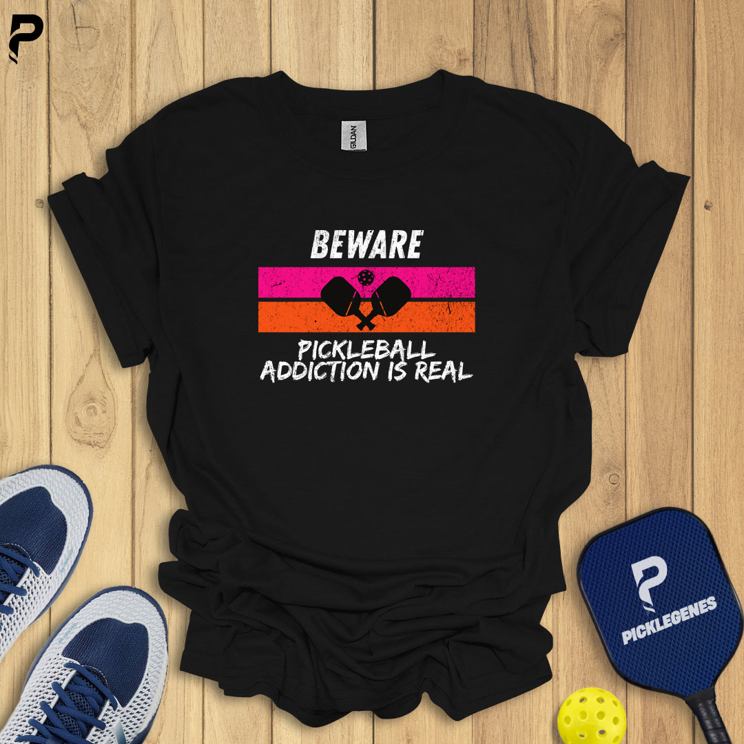 Black t-shirt with pickleball-themed text on a wooden surface with pickleball equipment.