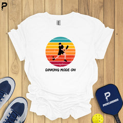 White t-shirt with pickleball design on a wooden surface with shoes and paddle.