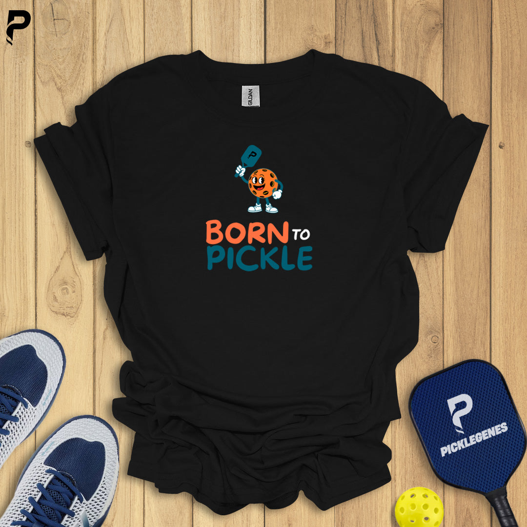 Black t-shirt with 'Born to Pickle' design on a wooden surface with pickleball equipment.