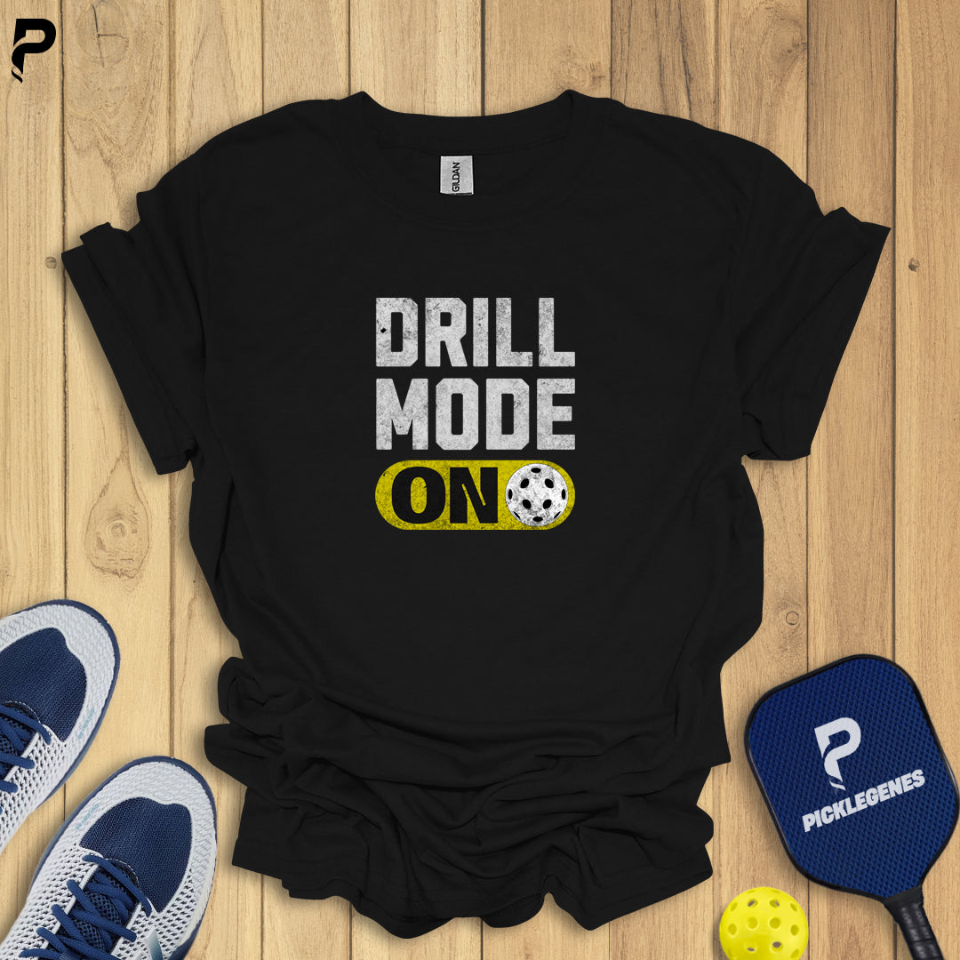 Black t-shirt with 'DRILL MODE ON' text on a wooden surface with sports equipment.