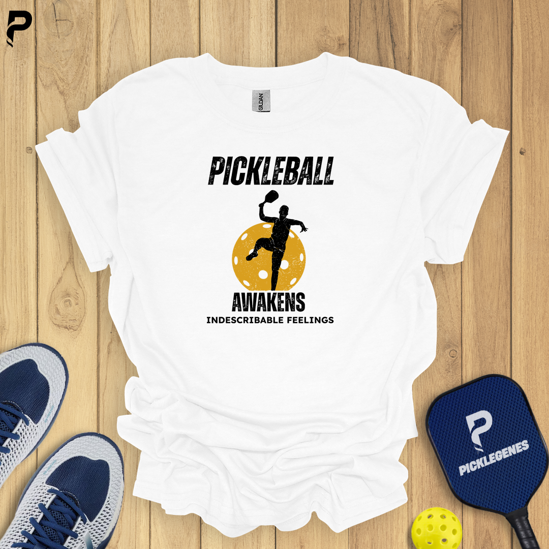 White t-shirt with pickleball-themed design on a wooden surface with pickleball equipment.